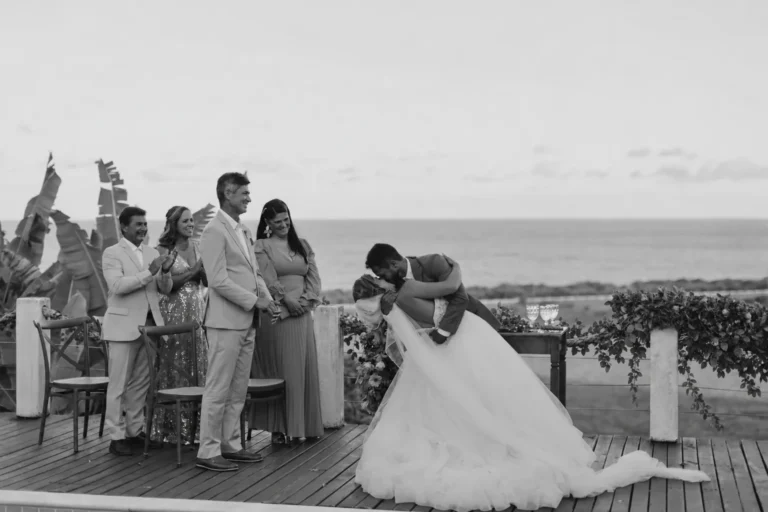 Beijo dos noivos no deck com os convidados comemorando e o mar ao fundo, em casamento no Azorai