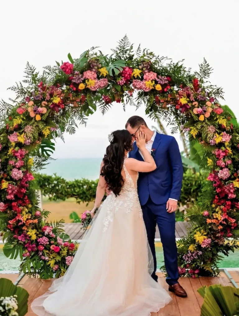 Beijo dos noivos sob arco de flores coloridas, com o mar ao fundo, em destination wedding no Azorai, Santa Cruz Cabrália