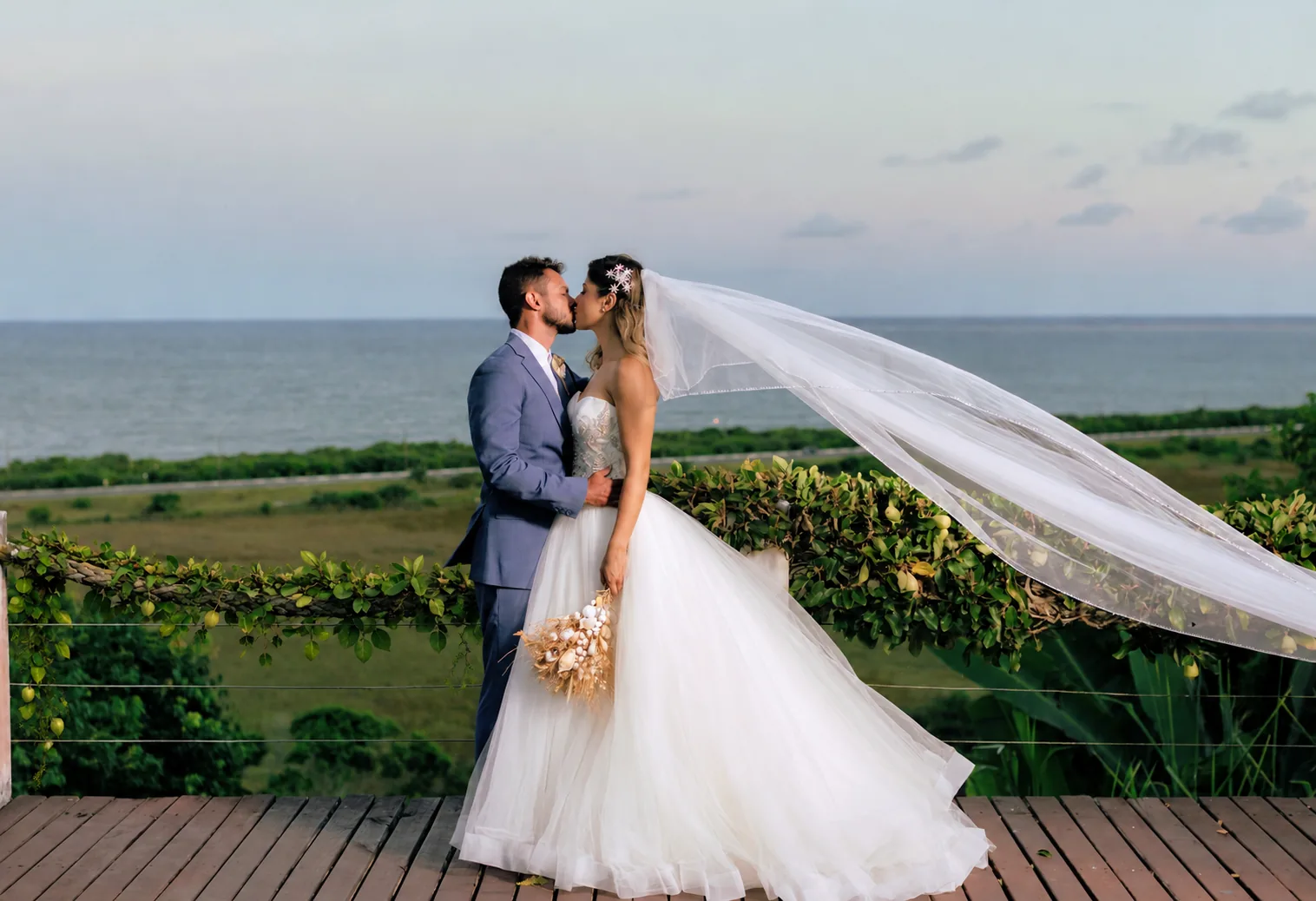 Noivos se beijam no deck com o véu ao vento e o mar ao fundo, em mini wedding no Azorai