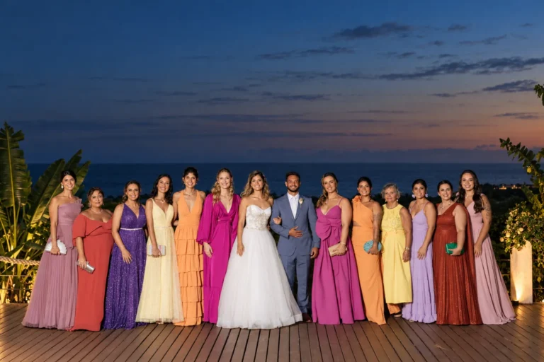 Noivos e madrinhas com vestidos coloridos no deck ao anoitecer, com o mar ao fundo, no Azorai